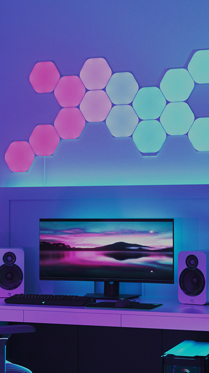 デスク上のモニターの上の壁のNanoleaf Shapes Hexagonsの画像です。 自宅環境でゲームを楽しむのに最適なRGB照明です。 色が変わる組み立て式ライトパネルはリンカーでつながり、1600万色以上のカラーバリエーションで光ります。