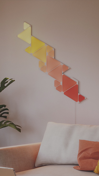リビングのソファの上の壁のNanoleaf Shapes TrianglesとMini Trianglesの画像です。 色が変わる組み立て式スマートライトパネルをリンカーで接続、1600万色以上のカラーバリエーション。